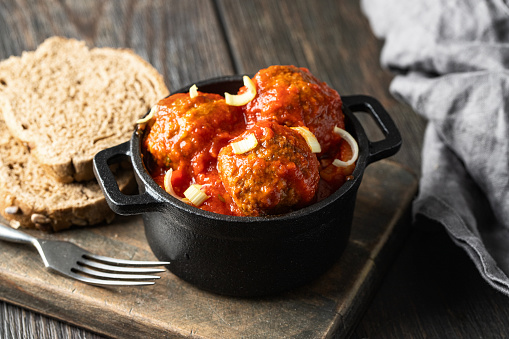 Iron mini pot with meatballs in tomato sauce on wooden cut board with vintage cutlery. Rustic style background