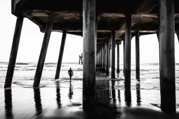 Monochromatic pier underside with a surfer walking into the sea