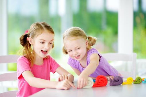 Two cute little sisters having fun together with colorful modeling clay at a daycare. Creative kids molding at home. Children play with plasticine or dough.