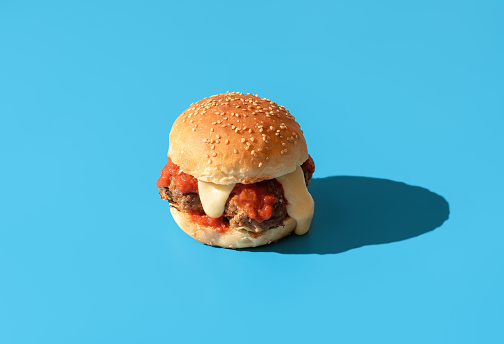 Homemade burger with meatballs, tomato sauce, and white cheddar sauce. Delicious burger in bright light minimalist on a blue table.