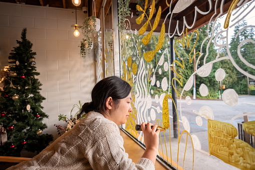 Artist using gold and white paint pens to decorate a shop window for the winter holiday season.  Vancouver, British Columbia, Canada.