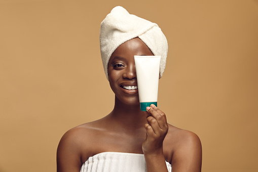 Happy young black skin woman holding jar of cream wearing in spa towel and head turban over beige background. Beauty, cosmetics and skincare concept.