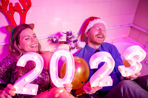 Cheerful young couple having fun at New Year's Eve party, holding illuminative numbers 2023 while sitting in a bathtub filled with balloons and confetti