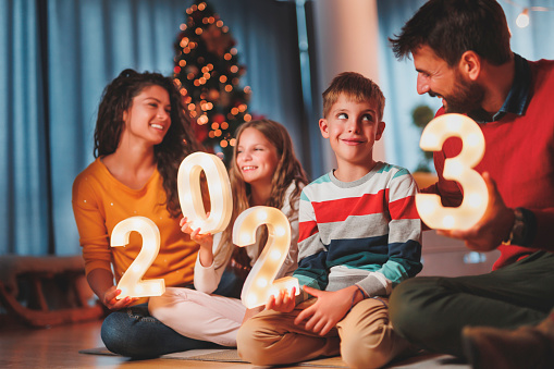 Parents celebrating New Years Eve at home with kids, sitting by the Christmas tree, holding illuminative numbers 2023 representing the upcoming New Year