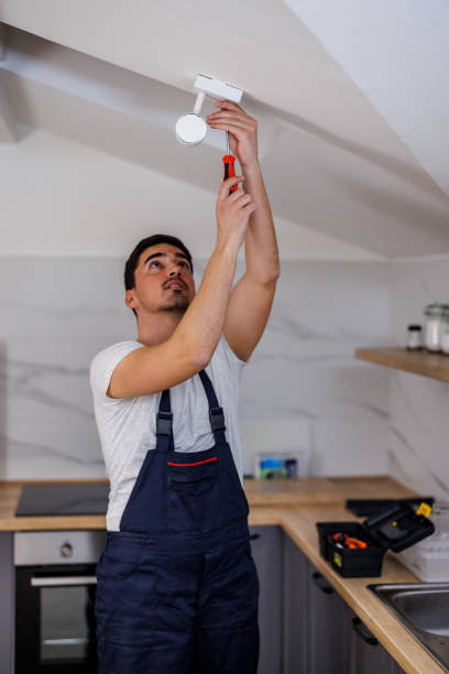 A young maintenance worker is fixing a fire alarm on a ceiling.