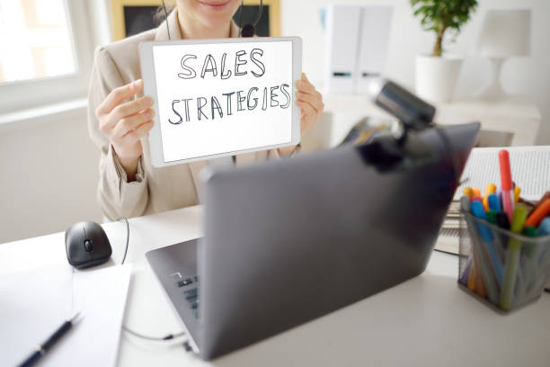Young woman is teaching english for business with laptop computer, camcorder and headphone at home. Teacher is having video conference chat with student. Person hold banner "Sales strategies" stock photo