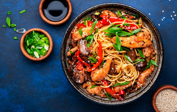 Stir fry noodles with chicken, red paprika, mushrooms, chives and sesame seeds in bowl. Asian cuisine dish. Blue table background, top view stock photo