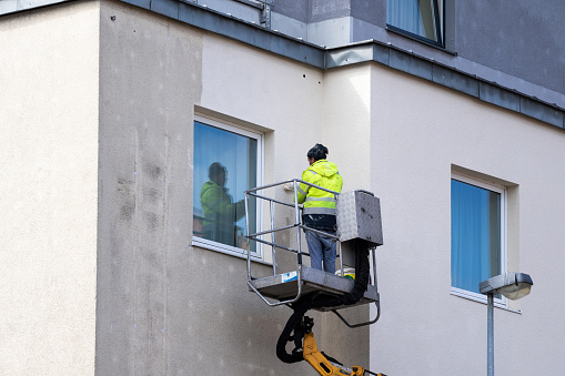 Maintenance worker in workwear using roller brush paints the facade of a residential house. Man in overalls standing outside building and repainting concrete wall close to window. Improvement concepts