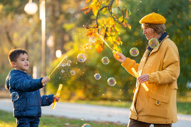 Active senior woman in yellow and her grandson playing with soap bubbles in the sunny autumn park. stock photo