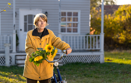 Smiling senior woman in yellow, carrying her bicycle in front of the house and holding a bouquet of sunflowers