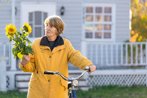 Smiling senior woman in yellow, carrying her bicycle in front of the house and holding a bouquet of sunflowers