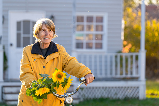 Smiling senior woman in yellow, carrying her bicycle in front of the house and holding a bouquet of sunflowers