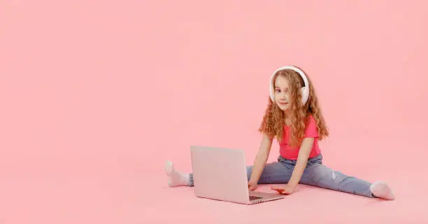 adorable child girl is sitting in the twine next to a laptop. kid in white headphones stretching and learns homework or watching cartoons, listening music.