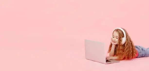 banner of adorable child girl is sitting in the twine next to a laptop. kid in white headphones stretching and learns homework or watching cartoons, listening music.