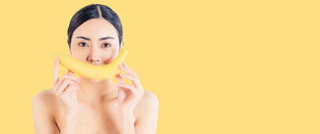 Beautiful woman holding banana and cover banana on her mouth It’s like a smiling face isolated on yellow background and copy space Beauty asian girl bear shoulder Diet female has healthy skin