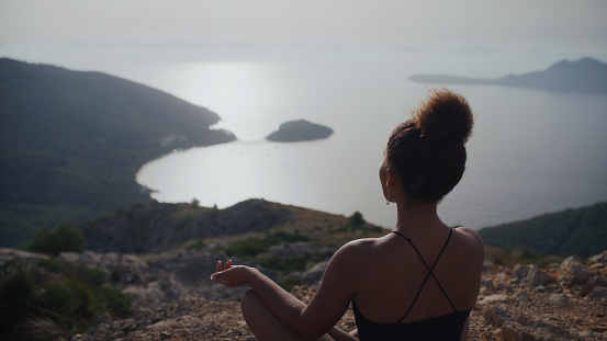 African ethnicity woman meditating in lotus position on top of a cliff and mountain looking at amazing view below