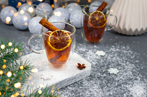 Two glass cups of tea with lemon and cinnamon sticks on the background of Christmas garlands and a gift