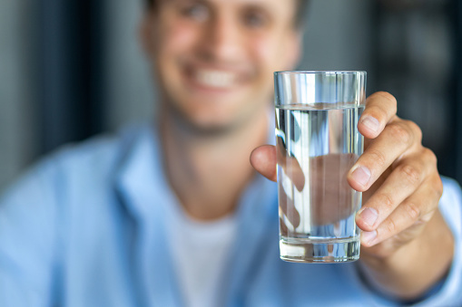 Young happy man hand, holds glass of fresh clear water give to camera and smiling, copy space