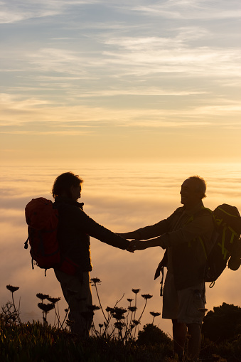 Dreamy silhouetted hikers at sunset. Man and woman in casual clothes and with ammunition standing at peak, holding hands. Hobby, active lifestyle, love concept