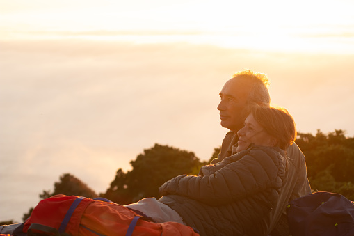 Happy elderly hikers at sunset. Man and woman in casual clothes and with ammunition sitting at peak, hugging. Hobby, active lifestyle, love concept