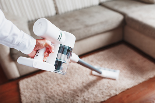 Powerful cordless vacuum cleaner with white cyclonic dust collection technology in hand, cleans the carpet in the house near the sofa. Close-up
