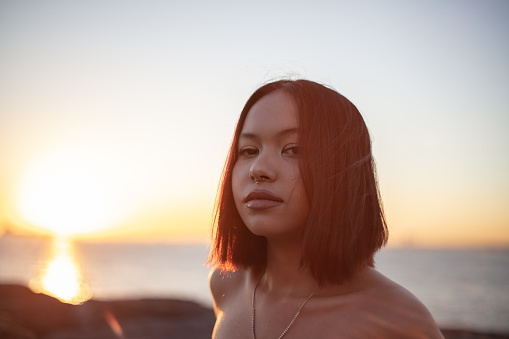 Portrait of beautiful asian girl. She is looking at the camera over by the sea at sunset time.