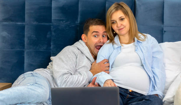 istock White Caucasian scared man, pregnant woman using, watching computer, laptop, sitting, lying on bed. Family, couple is preparing to baby delivery. Male wears in blue jeans and hoodie, female in shirt 1445018271