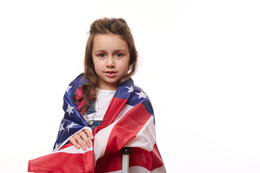 Happy preschooler girl wrapping in USA flag, smiling, posing with suitcase, isolated on white background, copy ad space. Kid travelling to America.Winning green card lottery. Citizenships. Emigration