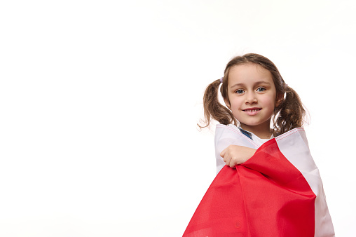 European little girl, proudly wrapping in flag of Poland or Canada, isolated on white background. Concept of national holidays. Patriotic events. Citizenships. Patriotism. Independence Day. Ad space