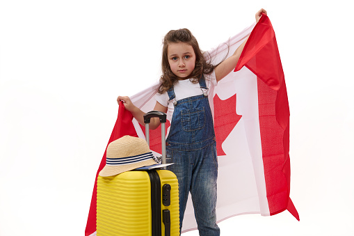 Cute little girl in white t-shirt and blue denim overalls, carrying Canada flag, smiles looking at camera while posing with a yellow suitcase over white background with free space for advertising text