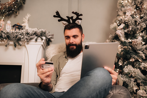 Young man doing some last minute Christmas shopping online. Man working during the holidays on his digital tablet, while sitting in his decorated living room, next to the fireplace.