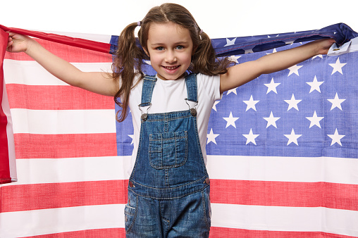 Happy Caucasian child, cute baby girl wearing blue denim overalls, smiling a beautiful toothy smile, looking at camera, holding American flag in her outstretched hands. 4th July - Independence Day