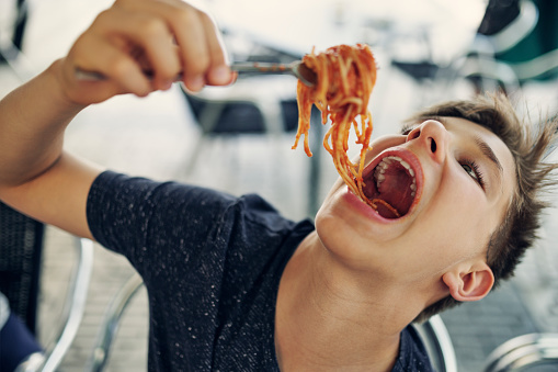 Teenage boy enjoying a spaghetti lunch. Teenage boy is biting on a fork full of spaghetti.\n\nCanon R5