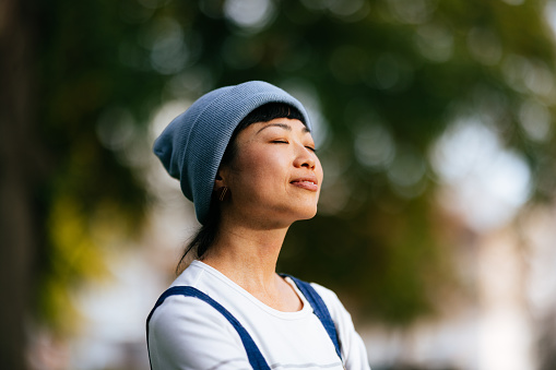 Close up photo of smiling Asian woman with closed eyes practicing deep breathing outdoors.
