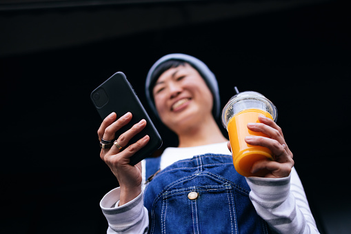 Low angle view of an anonymous Asian woman holding disposable cup with fresh citrus juice and typing text message on her smartphone while standing in front of black wall.