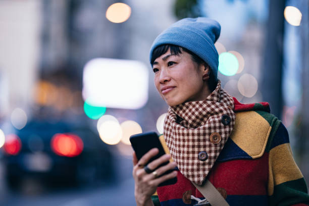 Happy Asian woman holding her smartphone while waiting for a taxi at the city street.