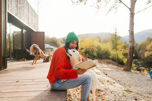 Photo of a young woman petting her dog while reading a book and enjoying her afternoon outside her remote house.