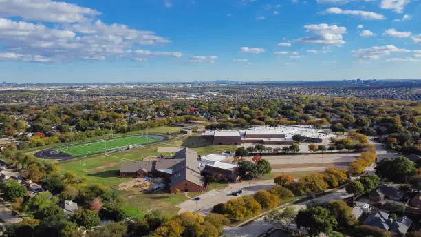Aerial view school district with football field, elementary and middle schools in upscale residential neighborhood with downtown Dallas and Las Colinas in distance background. Beautiful autumn leaves