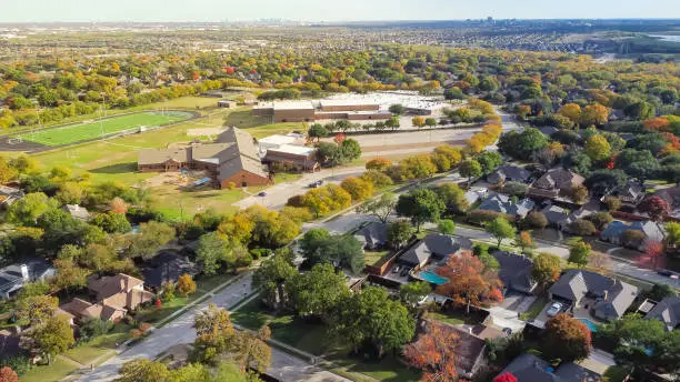 Aerial view school district with football field, elementary and middle schools in upscale residential neighborhood with downtown Dallas and Las Colinas in distance background. Beautiful autumn leaves