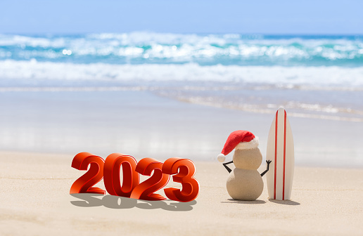 Sandy Christmas Snowman is watching the waves, standing on beautiful beach with a surf board. 2023 year concept