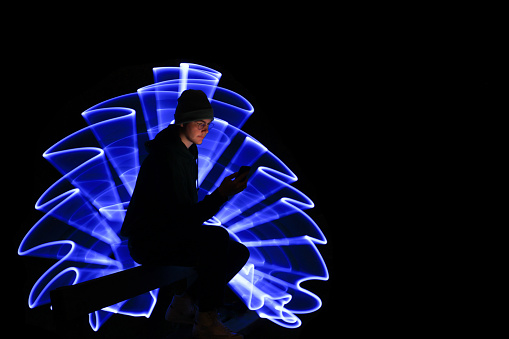 A young man sitting on a bench watching and using his mobile phone. Lightpainting scene in the background with blue light.