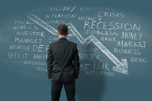 Economic crisis and related business terms word cloud written with chalk on a blue wall. There is a big arrow chart moving down. A businessman seen from the back is looking at the words