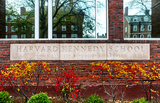 Cambridge, Massachusetts, USA - November 27, 2022:  Sign for the Harvard Kennedy School (HKS), officially the John F. Kennedy School of Government. It is the school of public policy and government of Harvard University in Cambridge. The school offers master's degrees in public policy, public administration, and international development, four doctoral degrees, and many executive education programs. Window reflections are of Harvard University campus buildings across JFK Street.
