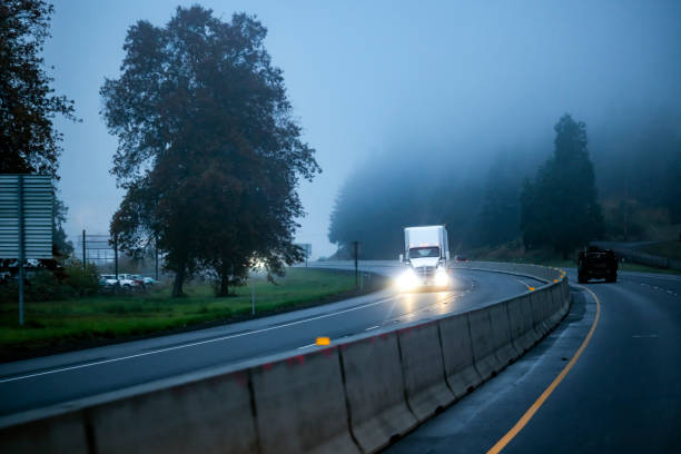istock Big rig white semi truck with turned on headlights transporting cargo in semi trailer running on the road with fog at the morning 1445073906