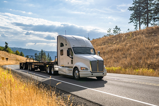 Industrial white long haul big rig high extended cab semi truck tractor transporting empty flat bed semi trailer running on the narrow winding highway road to warehouse for the next load