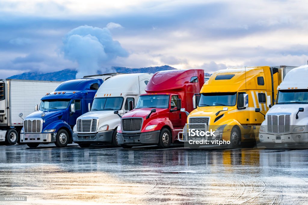 Bright colorful big rigs semi trucks with semi trailers standing in the row on truck stop parking lot at early morning Different make big rigs semi trucks tractors with loaded semi trailers standing in the row on truck stop parking lot at early morning waiting for the route continuation time according to the log book Truck Stock Photo
