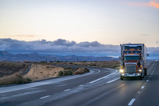 istock Brown classic big rig semi truck tractor with turned on headlight transporting cargo in reefer semi trailer driving on the straight one way road with fork intersection at twilight 1445075714