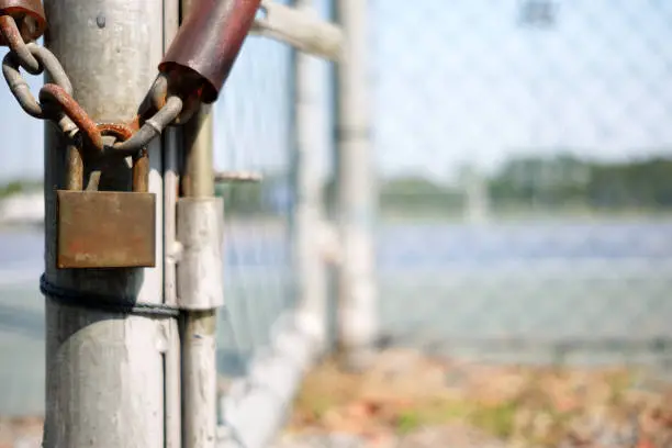 Photo of Old Rusty Padlock Hanging on Chain with Blurred Metal Fence Background.