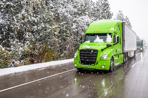 Bright green big rig semi truck transporting cargo in refrigerator semi trailer driving in front of convoy moving slowly on winter highway during a snow storm in the Lake Shasta region of California