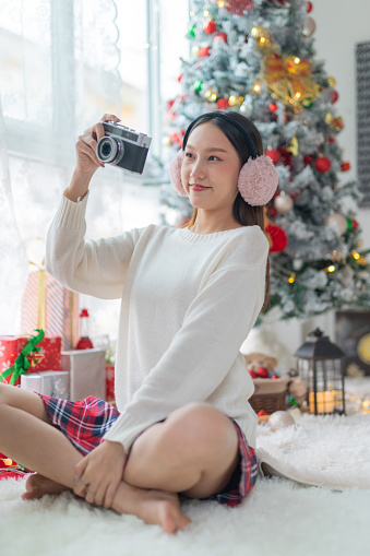 Portrait photo of a young beautiful friendly asian female lady with winter ear muffs cheerfully snapping photos with a vintage film camera in front a nice decorated christmas tree in her room